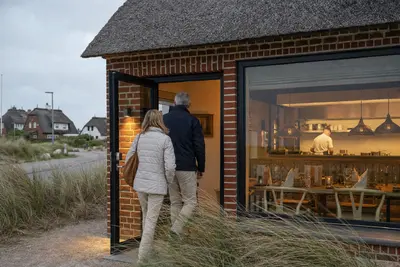 Paar betritt ein reetgedecktes Backsteinhaus hinter Dünengras, durchs Fenster sind Küche, Koch und gedeckte Tische zu sehen