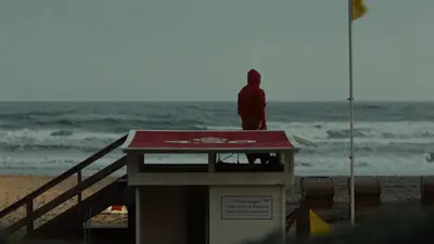 Wenningstedt: Person in roter Kapuze auf einem Strandhäuschen blickt aufs graue Meer, rechts weht eine gelbe Flagge
